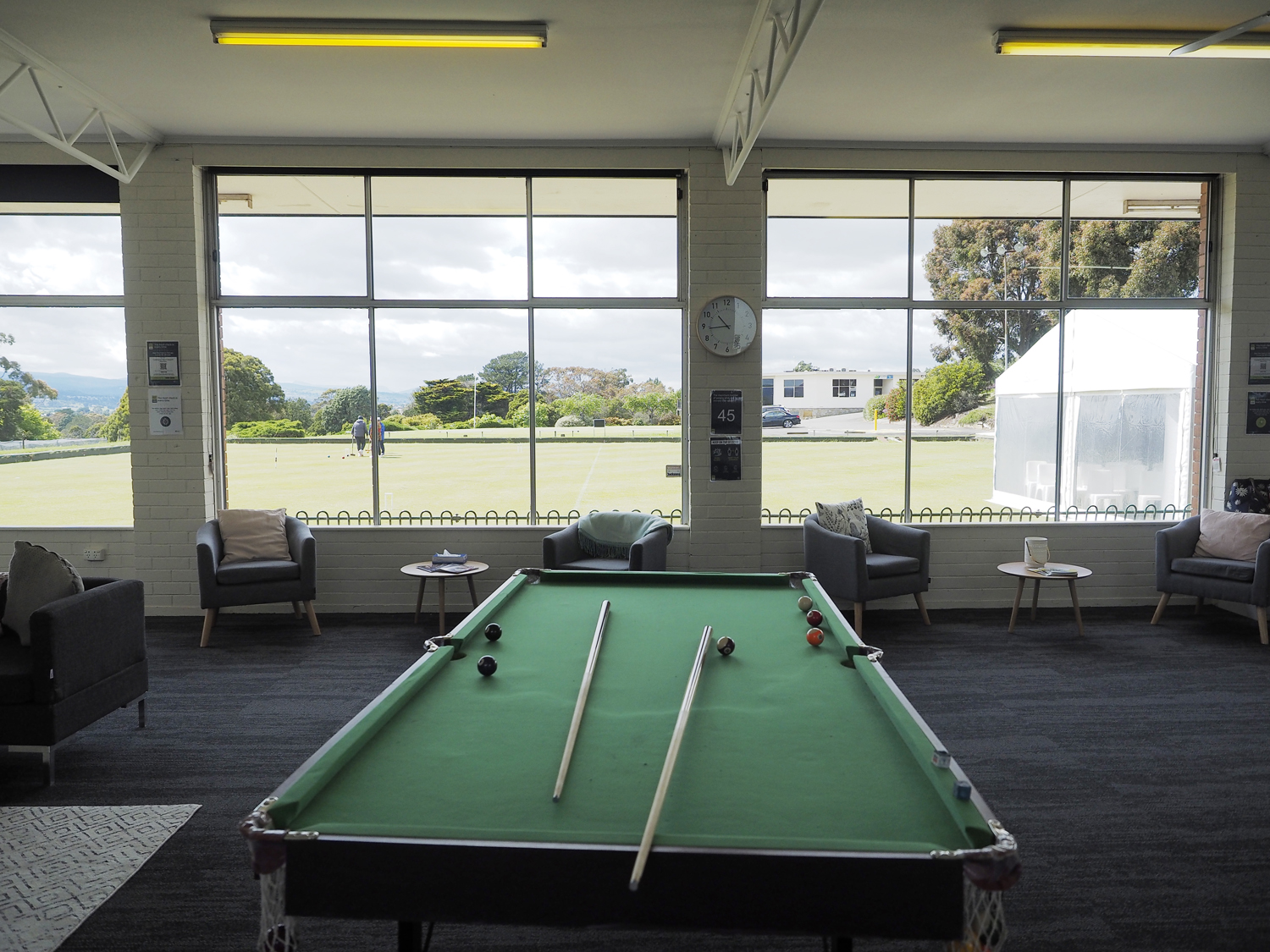 A pool table with cues in front of large windows looking out onto a lawn bowls green