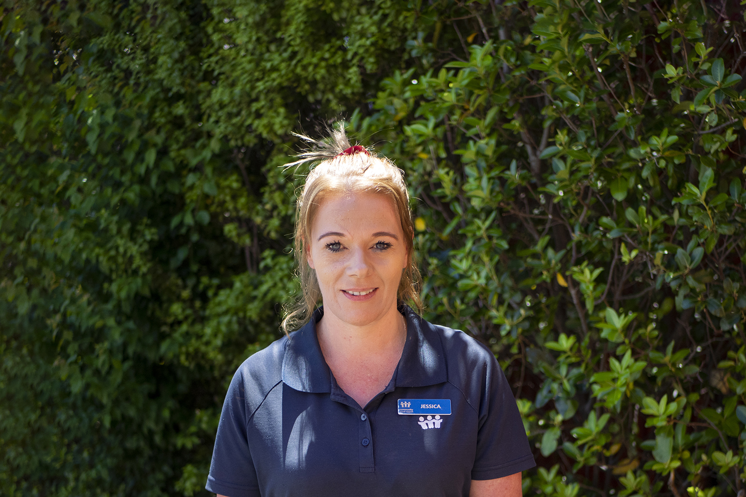 A woman in blue polo shirt and name tag stands for a photo in front of a green hedge