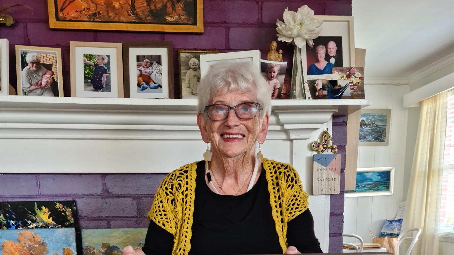 A lady with black top and yellow cardigan wearing glasses poses for a photo in her living room