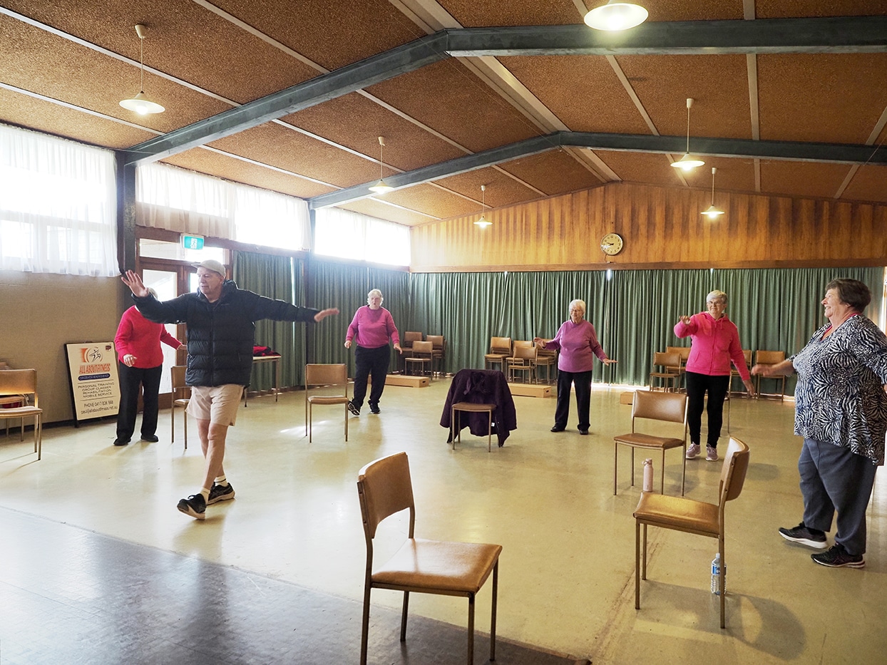 An exercise class for seniors taking place inside a hall with wooden panels and green curtained walls. There is a man with a cap, black puffer jacket and shorts leading the class of five women standing up and exercising.