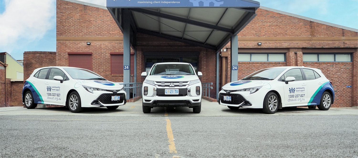 Three white vehicles with blue and green branding parked in front of a large red bricked building. The cars have the Community Based Support blue logo with three people and contact information.