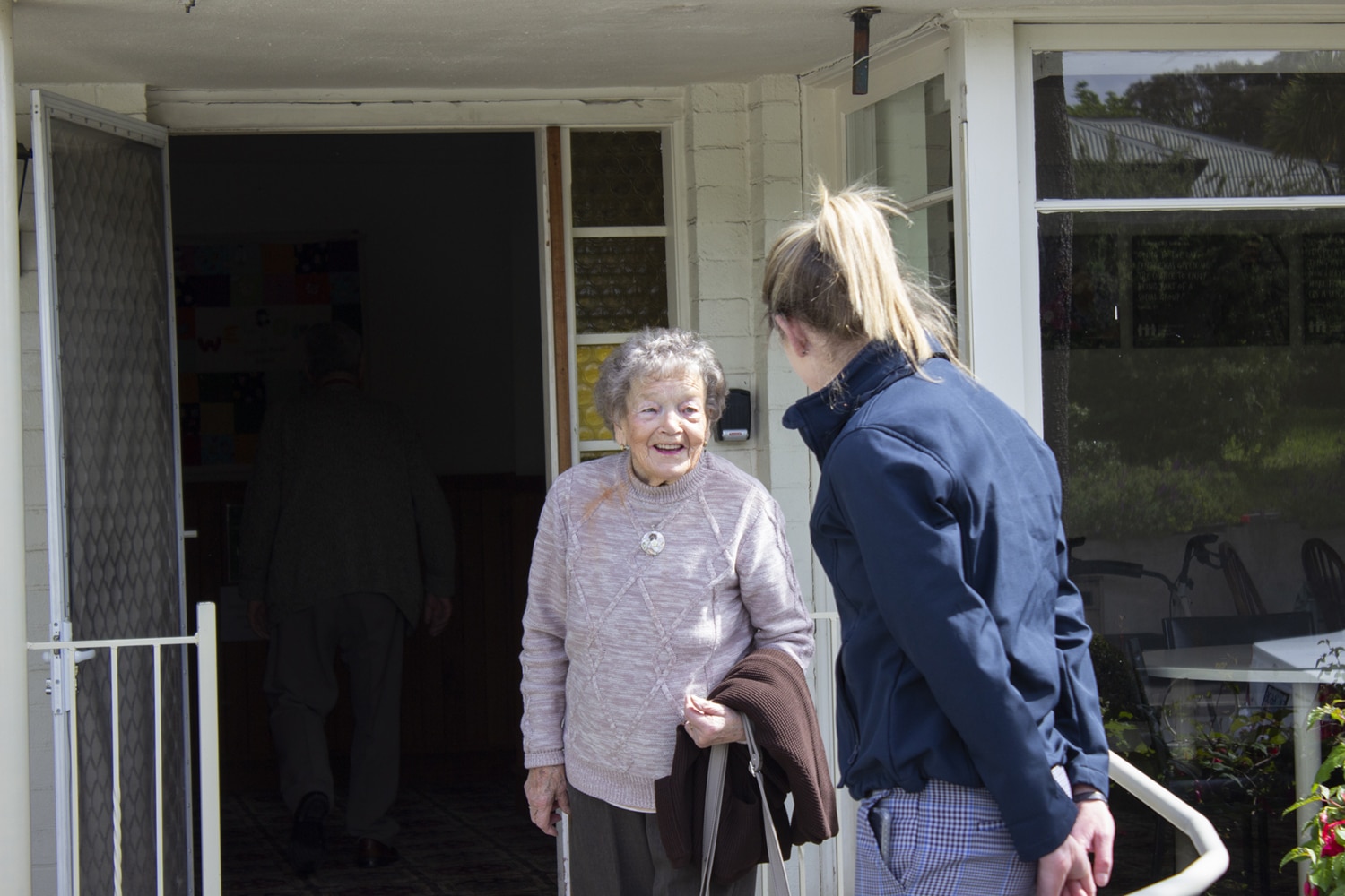 An older lady in a lavender sweater talks with a support worker in a blue jackout outside a social group venue.