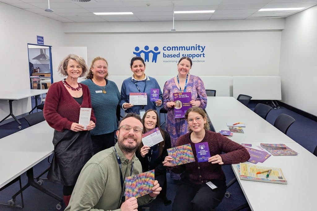 A group of seven smiling people standing and sitting in a meeting room at Community Based Support, holding colourful brochures and information sheets about elder abuse awareness.