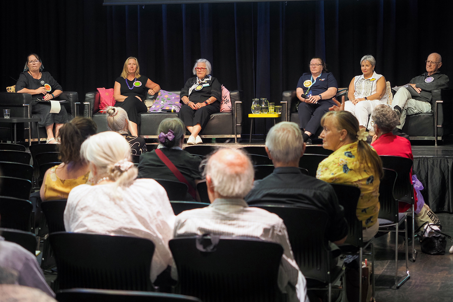 A panel of seven speakers seated on stage in front of a dark curtain, taking part in a discussion at the Brain Health Expo in Glenorchy. An audience is seated in the foreground listening, with some attendees turned towards the stage.