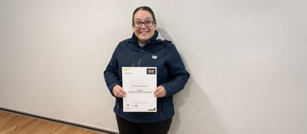 Iolanda Gregorio smiling and holding her Certificate III in Individual Support in front of a light wall.