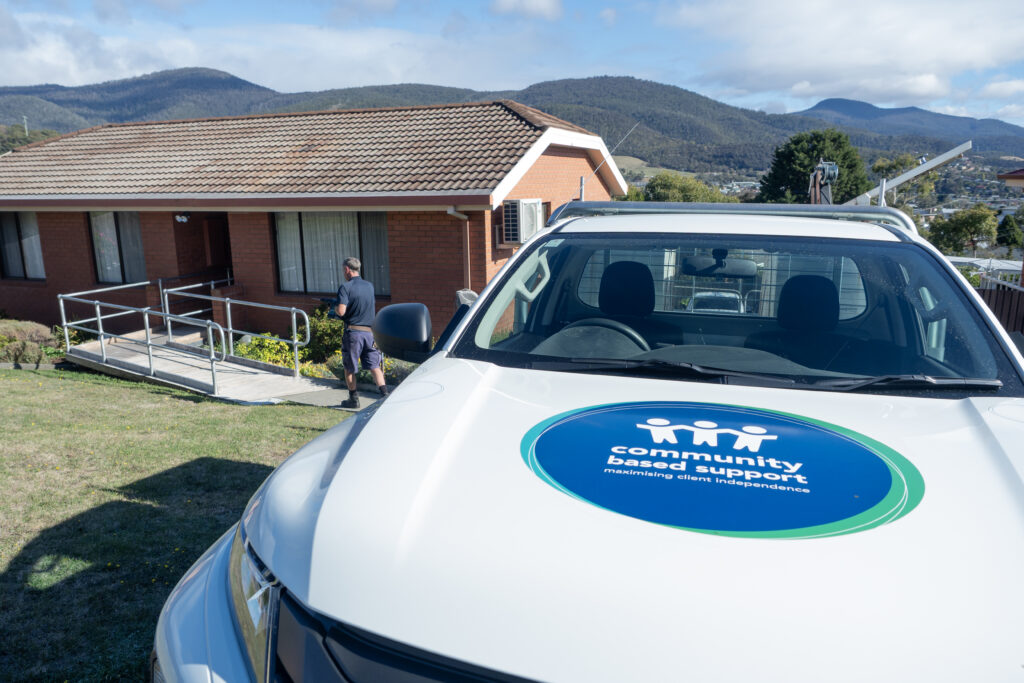 A Community Based Support vehicle parked outside a brick home with an accessibility ramp, as a staff member approaches the entrance.