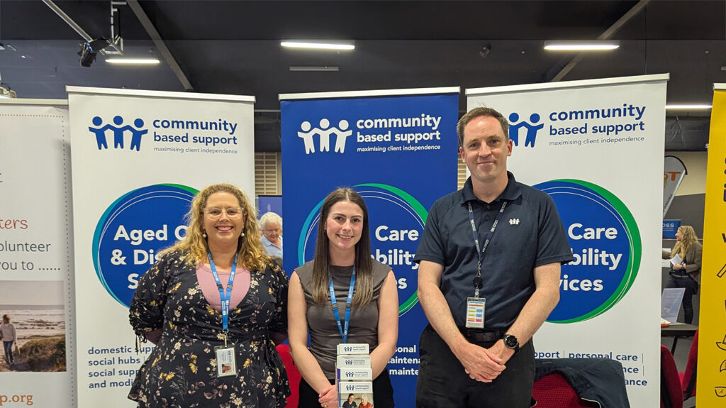 Three Community Based Support staff standing behind an information table at the Ageing Together Expo, with banners promoting aged care and disability services.